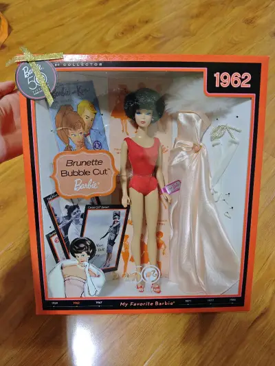 这是一款芭比50周年纪念版1962 Brunette Bubble Cut Barbie收藏级娃娃，它是芭比历史传承与时尚魅力的经典缩影。
 
1962年，“泡泡头”（Bubble Cut）发型的芭比横空出世，这款娃娃复刻了当年的经典造型。深棕色的泡泡头短发蓬松俏皮...