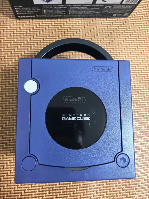 这是一台任天堂GameCube（NGC）家用游戏机，经典的紫罗兰紫配色。
机身上方是光盘仓，正面配备四个手柄接口，下方有SLOT‑A、SLOT‑B两个记忆卡插槽，右下角印有ATI的标识，代表该机的图形芯片由ATI提供。
 这台是任天堂2001年推出的第六代家用主机，...