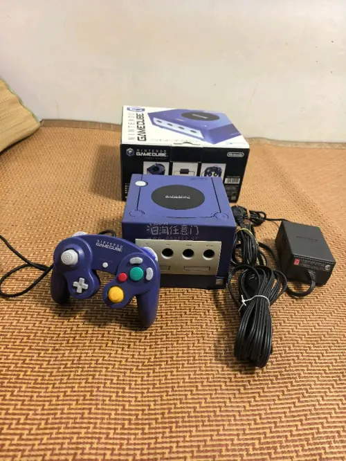 这是一台任天堂GameCube（NGC）家用游戏机，经典的紫罗兰紫配色。
机身上方是光盘仓，正面配备四个手柄接口，下方有SLOT‑A、SLOT‑B两个记忆卡插槽，右下角印有ATI的标识，代表该机的图形芯片由ATI提供。
 这台是任天堂2001年推出的第六代家用主机，...
